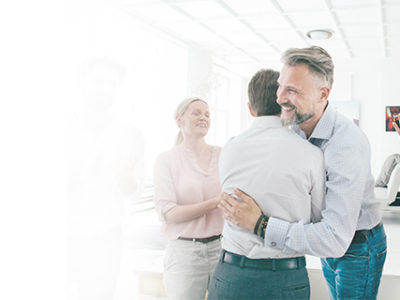 Two men hugging in the office with other people cheering