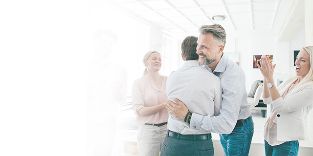 Two men hugging in the office with other people cheering