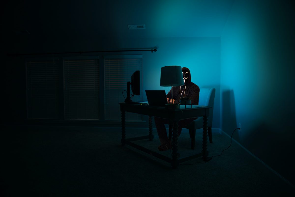 Hacker behind desktop computer in dark room, blue lit by screen | The Rise of Invoice Fraud in the Age of COVID