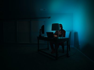 Hacker behind desktop computer in dark room, blue lit by screen | The Rise of Invoice Fraud in the Age of COVID
