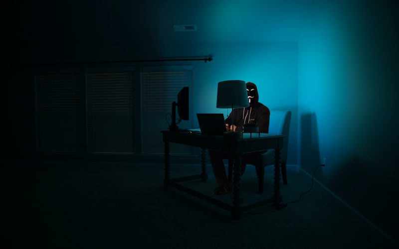 Hacker behind desktop computer in dark room, blue lit by screen | The Rise of Invoice Fraud in the Age of COVID